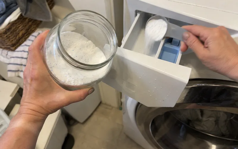 Hand adding one tablespoon of homemade detergent powder to an HE front-loader's detergent drawer