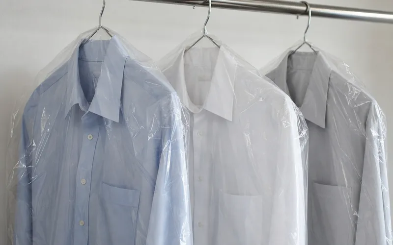 Pressed button-down shirts in clear plastic dry cleaning bags hanging on a metal closet rod