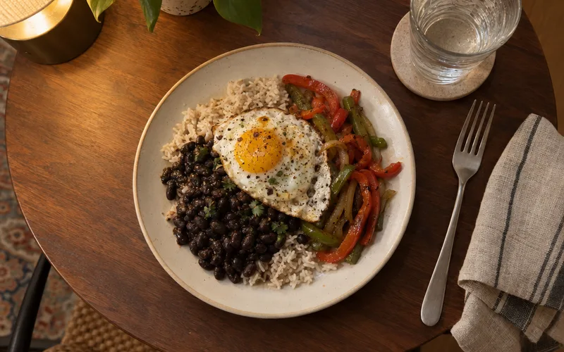 A single plate of rice, black beans, sauteed peppers and a fried egg on a small table set for one