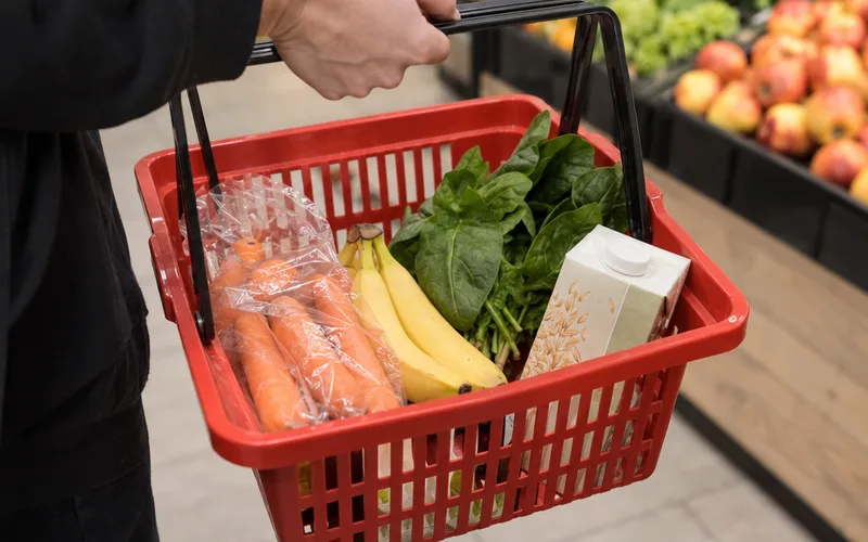A shopper's hand basket holding carrots, bananas, spinach and oats in a grocery store produce aisle